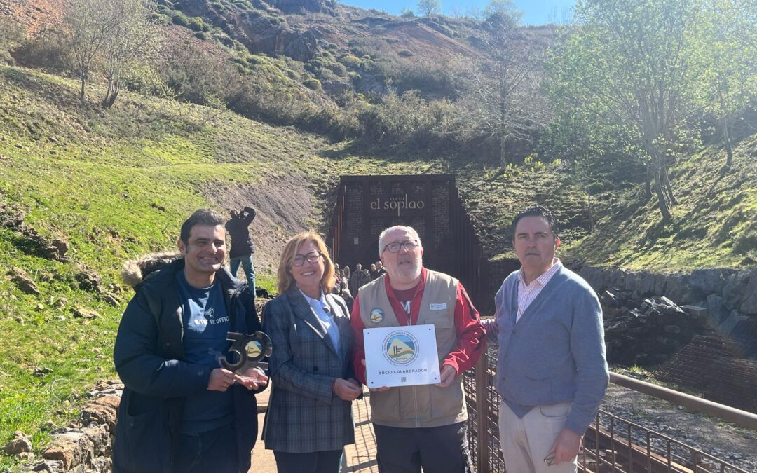 Cuatro personas posan en la entrada de la Cueva El Soplao, Cantabria. Dos de ellas sostienen una placa blanca que dice "Socio Colaborador" con el logotipo de la SEDPGYM. El fondo muestra una ladera verde y la estructura metálica de acceso a la cueva.